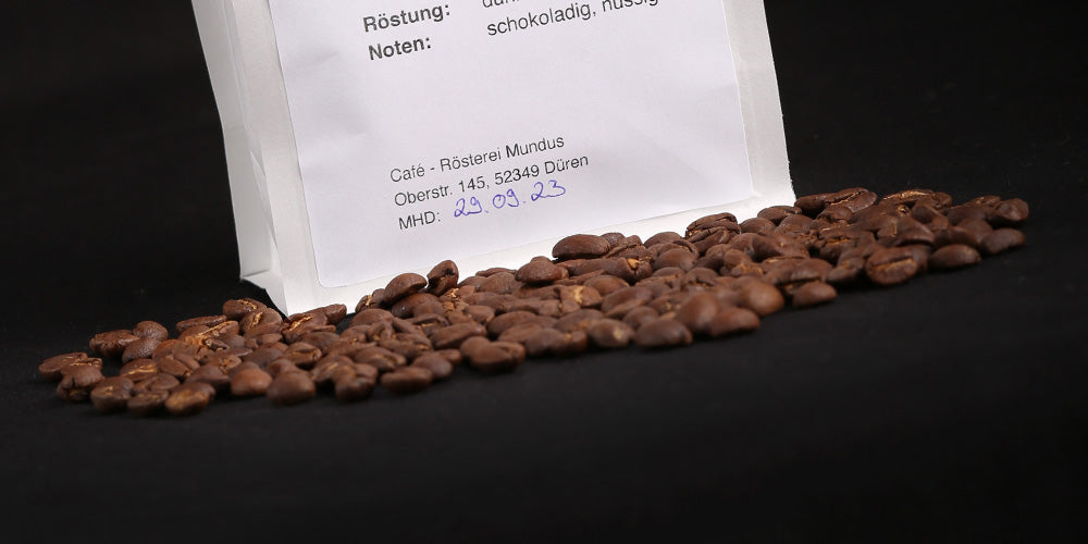 Eine weiße Kaffeetüte aus dem Café Mundus mit davor liegenden gerösteten Kaffeebohnen