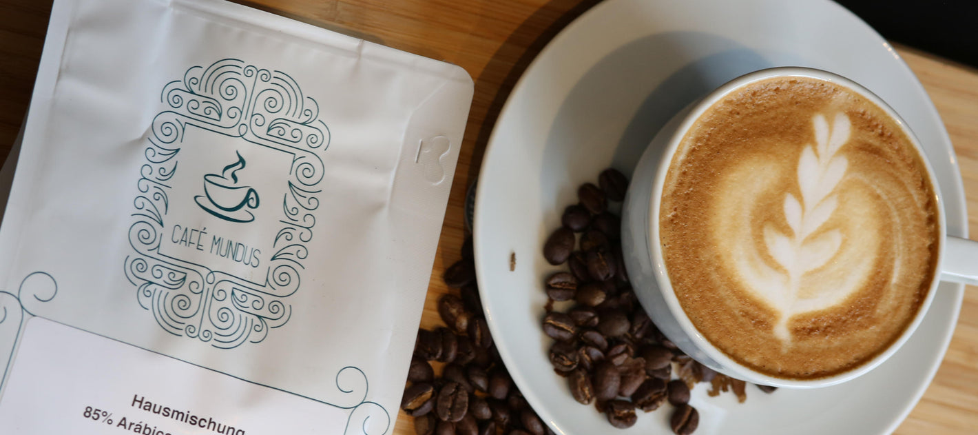 Eine 250g Kaffeetüte mit der Hausmischung aus der Rösterei Mundus neben einer Tasse Cappuccino mit einer Tulpe Latte Art und einigen dunklen Kaffeebohnen