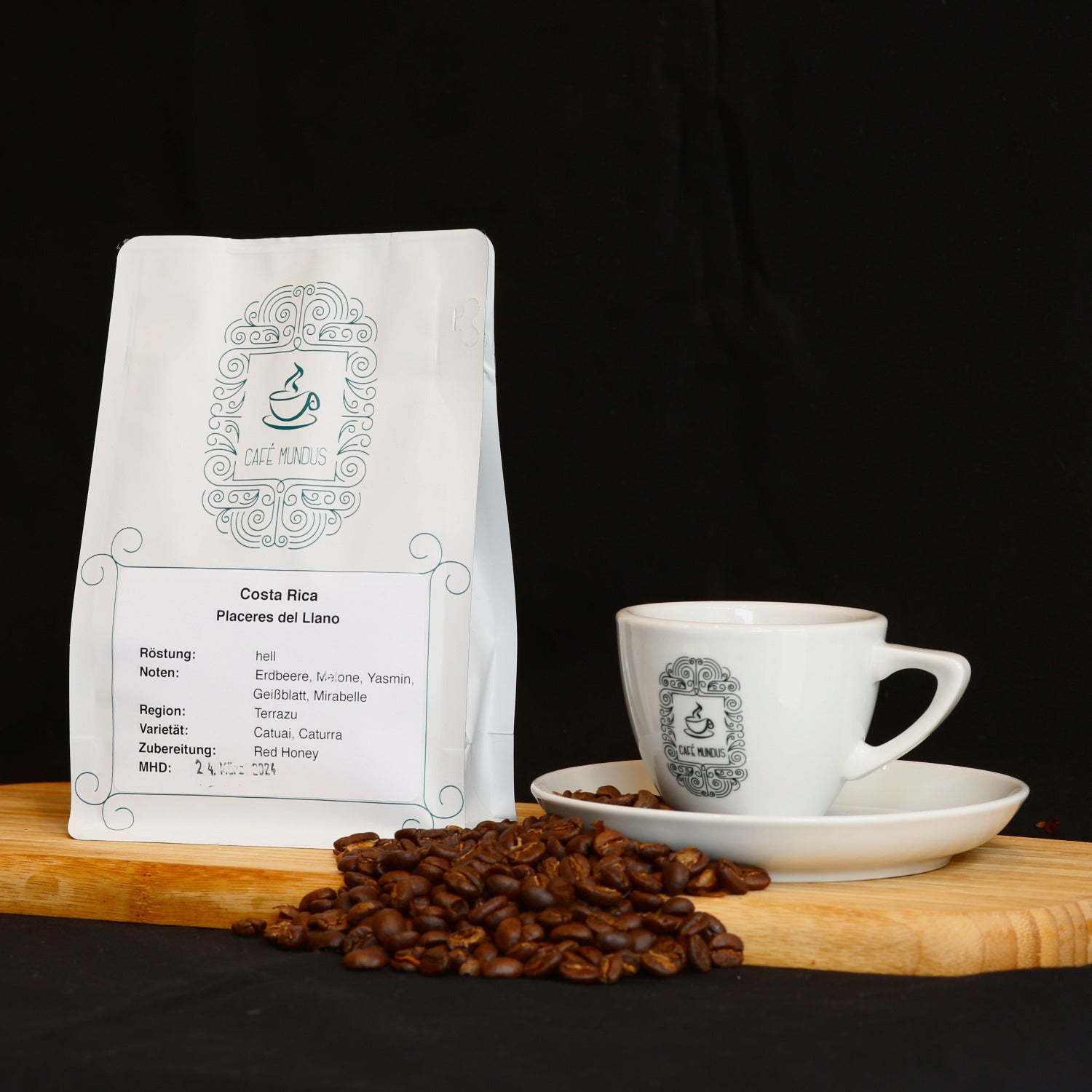 Eine weiße 250g-Tüte Costa Rica Kaffee aus der Rösterei Mundus in Düren auf einem hölzernen Tablett neben einer Kaffeetasse aus dem zugehörigen Café und einigen Kaffeebohnen