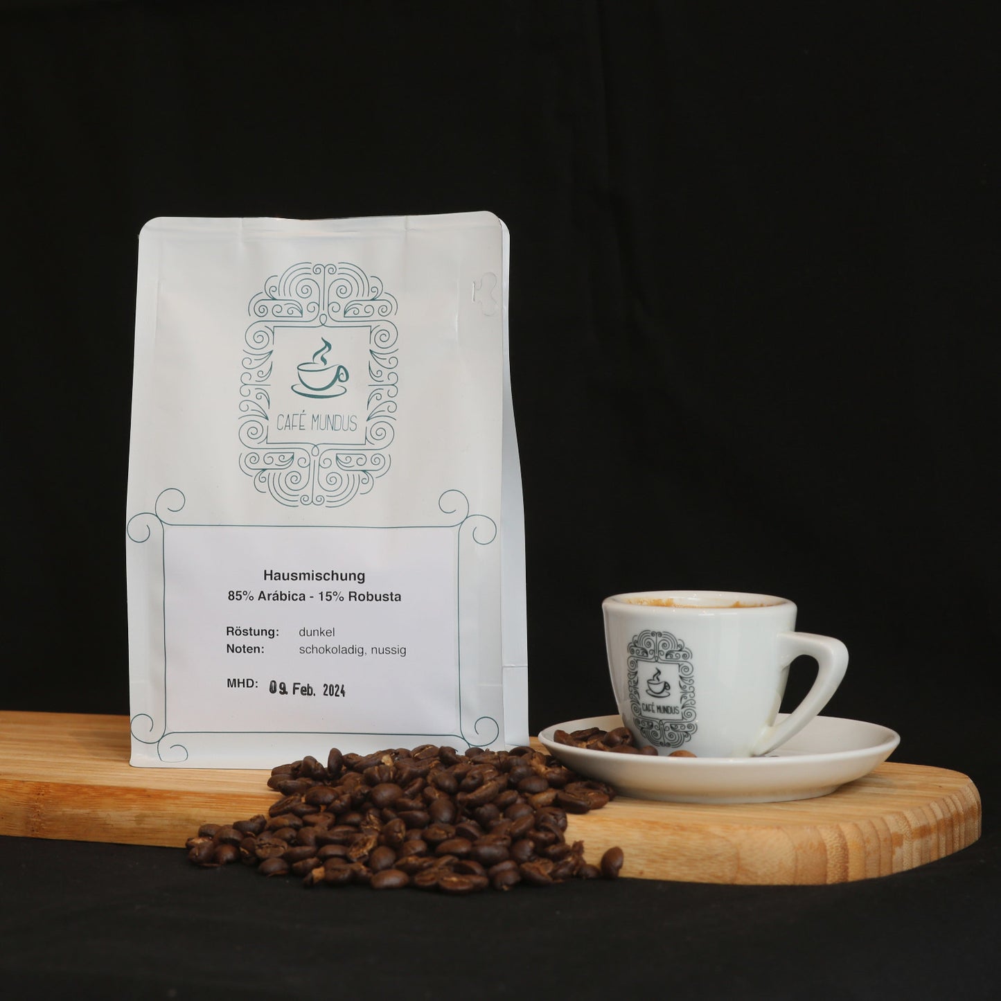 Eine 250g-Packung mit der Hausmischung aus der Rösterei Mundus auf einem Tablett neben einer Tasse Kaffee aus dem zugehörigen Café und einigen Kaffeebohnen