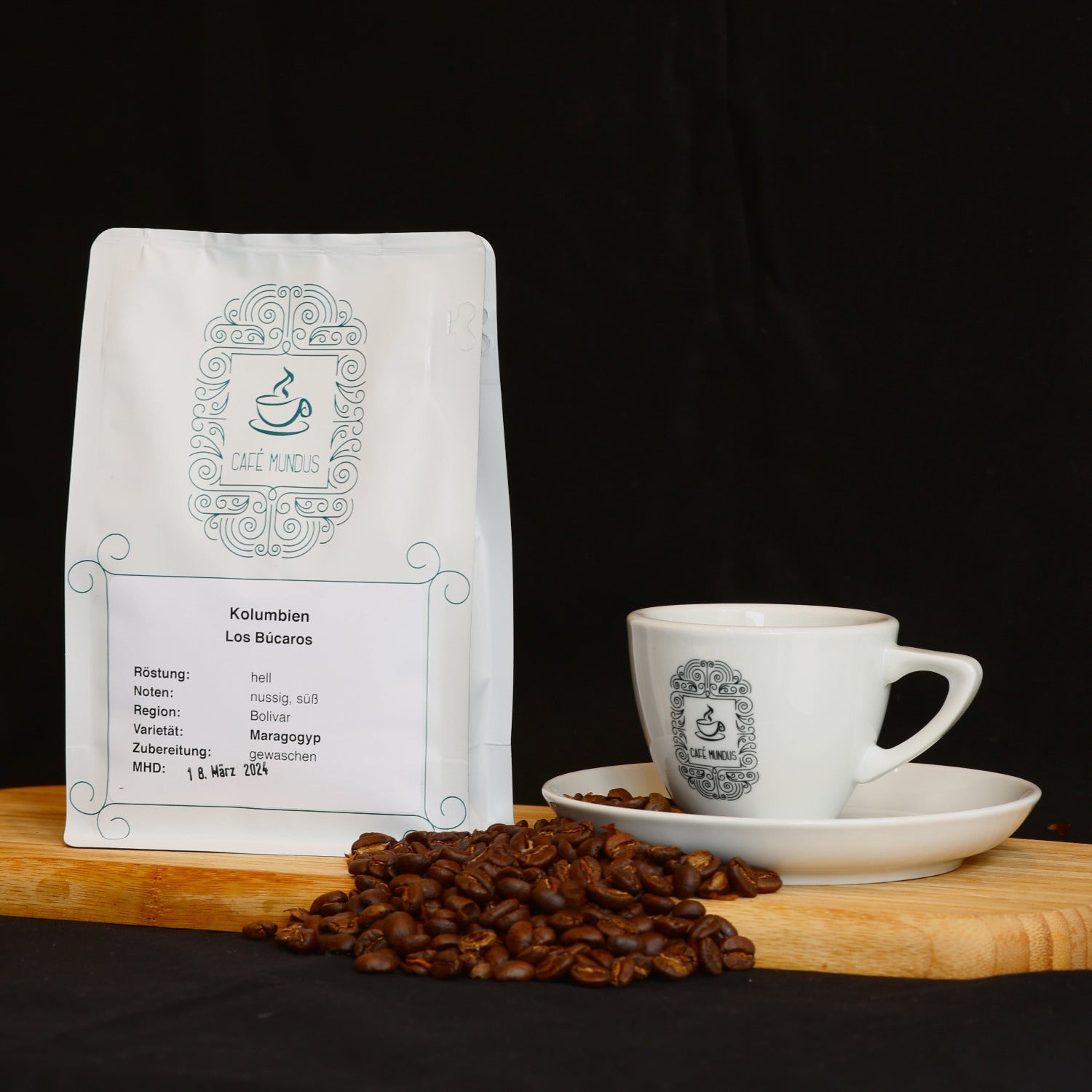 Eine weiße 250g Kaffeetüte mit der Kolumbien Los Bucaros Bohne aus der Rösterei Mundus in Düren auf einem hölzernen Tablett neben einer Kaffeetasse aus dem zugehörigen Café und einigen Kaffeebohnen